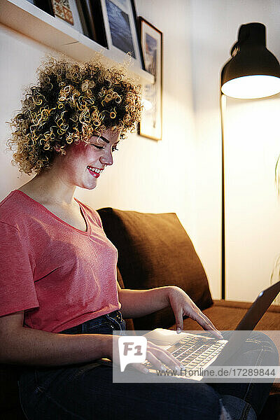 Lächelnde junge Frau beim Tippen mit dem Laptop im Wohnzimmer zu Hause