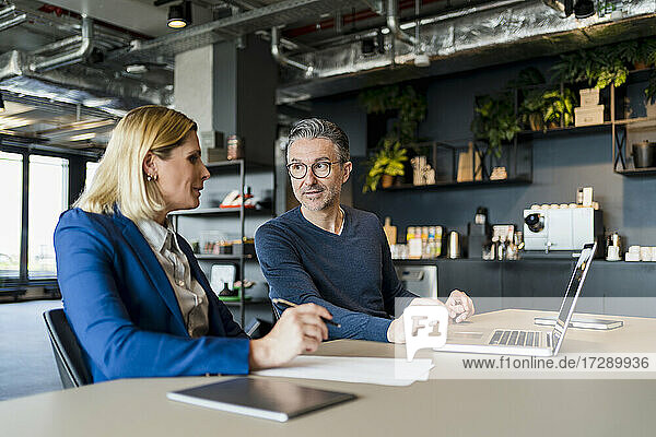 Männlicher Unternehmer im Gespräch mit einer weiblichen Fachkraft  die mit einem Laptop in einem kreativen Büro sitzt