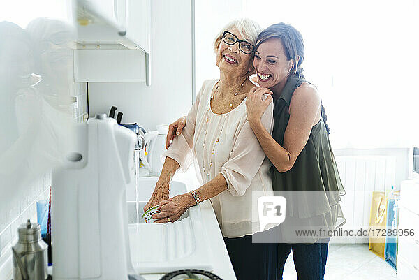 Fröhliche Enkelin umarmt Großmutter beim Geschirrspülen in der Küche zu Hause