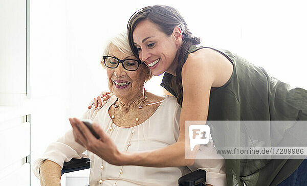Lächelnde Frau  die ein Selfie mit ihrer behinderten Großmutter über ihr Smartphone zu Hause macht
