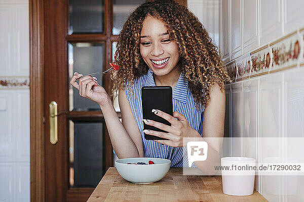 Lächelnde junge Frau benutzt ihr Smartphone beim Frühstück in der Küche