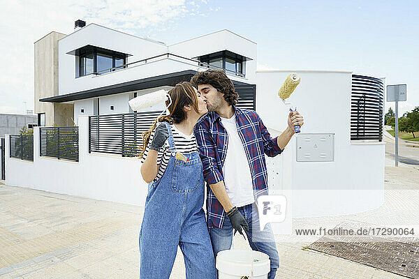 Pärchen mit Farbrolle küsst sich vor einem neuen Haus