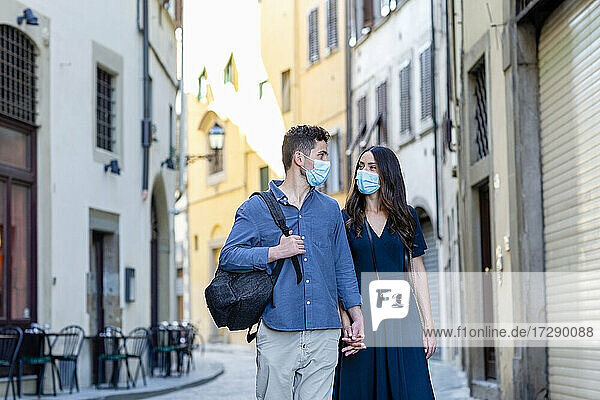 Touristenpaar mit Gesichtsschutzmaske  das sich auf einer Straße in der Stadt an den Händen hält