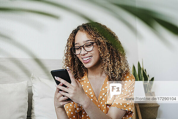 Lächelnde  lockig behaarte Frau  die über ihr Smartphone im Schlafzimmer zu Hause Textnachrichten verschickt