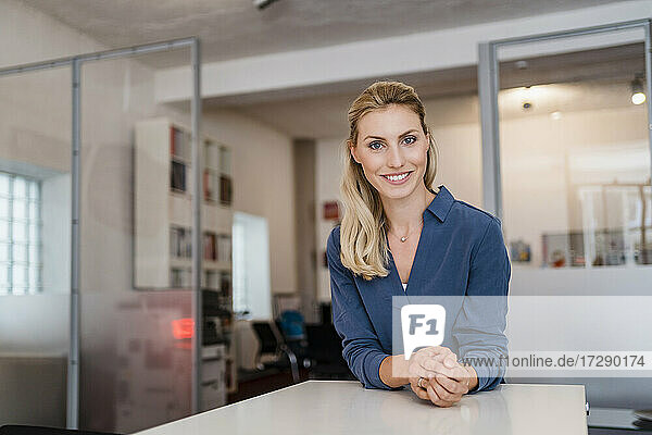 Lächelnde schöne blonde Geschäftsfrau lehnt sich an den Schreibtisch im Büro