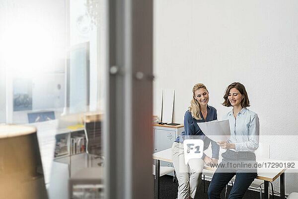 Glückliche junge Geschäftsfrau  die über ein Dokument diskutiert  während sie auf dem Schreibtisch im Büro sitzt