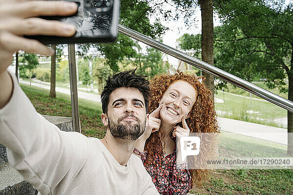Young couple making cute faces while talking selfie in public park