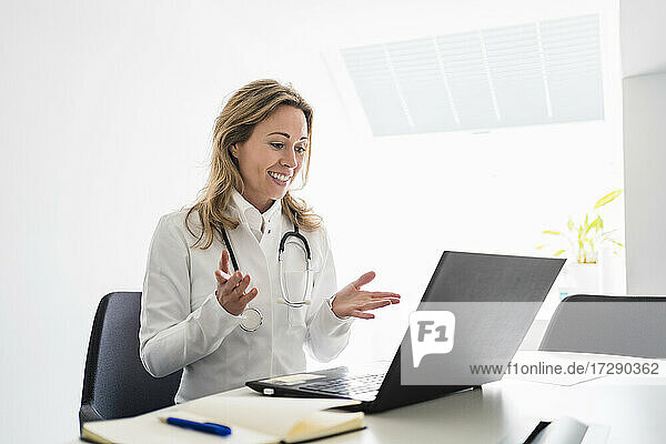 Lächelnde Ärztin gestikuliert während eines Videogesprächs über einen Laptop am Schreibtisch