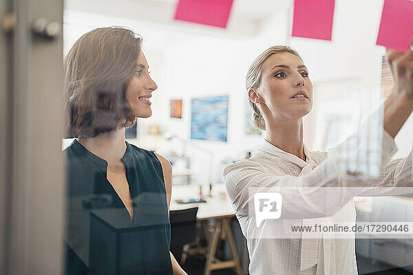 Lächelnde Geschäftsfrau  die eine Kollegin bei der Arbeit im Büro betrachtet  scheint durch Glas zu schauen