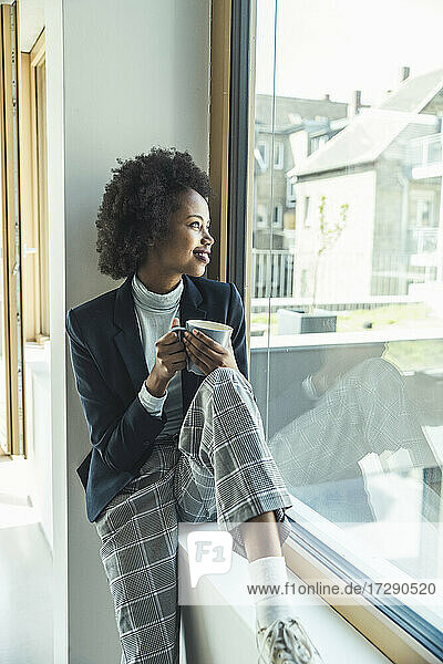 Lächelnde Geschäftsfrau  die auf der Fensterbank im Büro sitzt und träumt