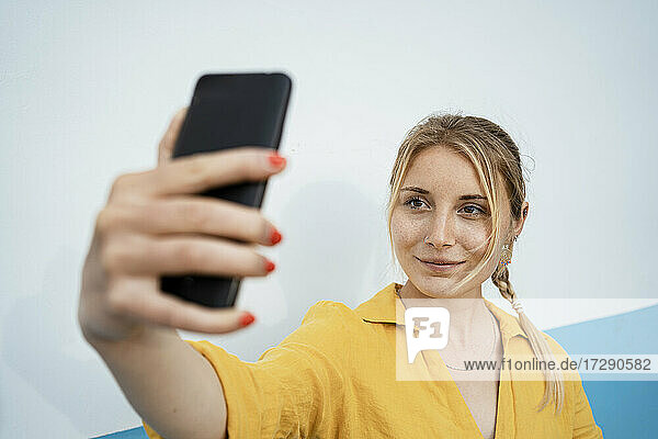 Lächelnde Frau nimmt Selfie durch Handy von weißen Wand