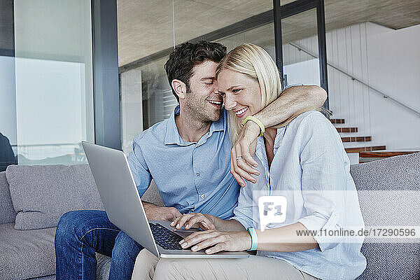 Lächelnder Mann umarmt Frau  die einen Laptop benutzt  während sie auf dem Sofa im Wohnzimmer sitzt