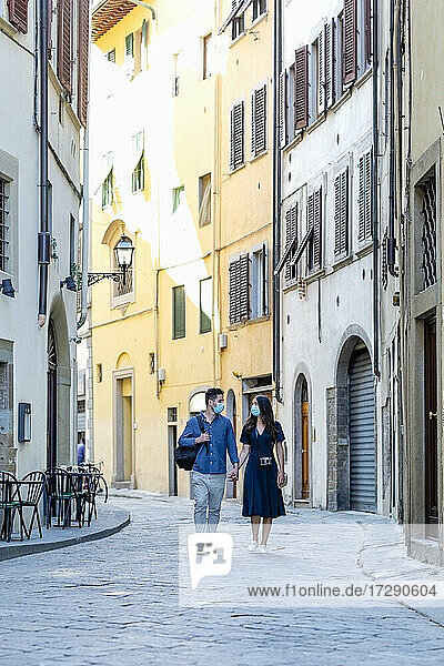 Touristisches Paar mit Gesichtsmaske  das sich beim Gehen auf einer Straße in der Stadt an den Händen hält