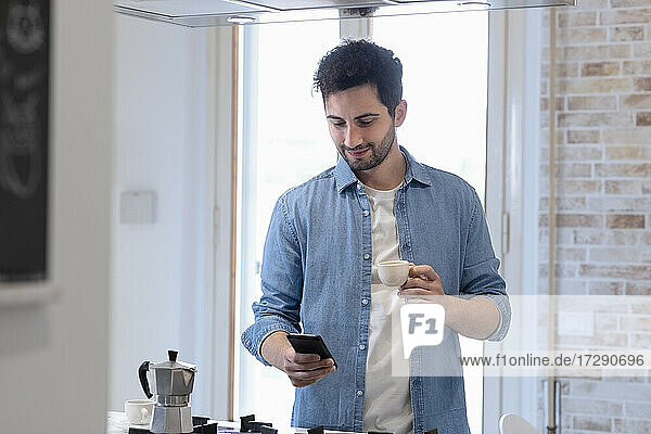 Young man using mobile phone while holding coffee cup in domestic kitchen