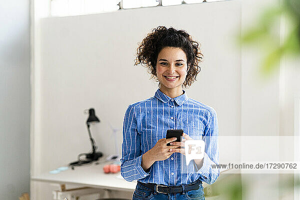 Lächelnde Geschäftsfrau  die ein Smartphone hält  während sie in einem kreativen Büro steht