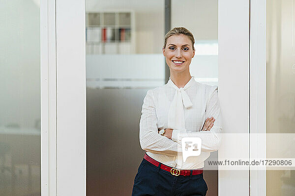Smiling female entrepreneur standing with arms crossed leaning at doorway in office