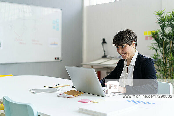 Lächelnde mittlere erwachsene Geschäftsfrau  die einen Laptop benutzt  während sie am Schreibtisch im Büro sitzt