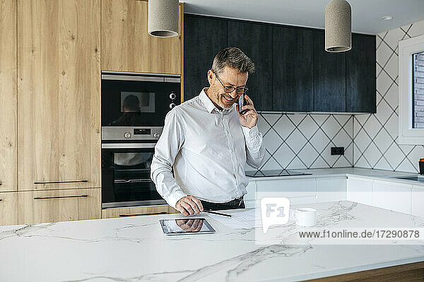 Lächelnder Geschäftsmann  der über sein Smartphone spricht  während er in der Küche zu Hause an einem Geschäftsplan arbeitet