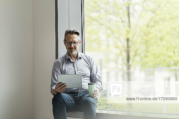 Geschäftsmann mit Einwegbecher und digitalem Tablet am Bürofenster