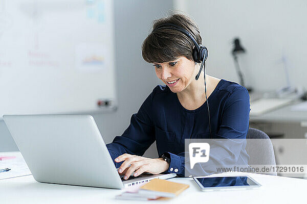 Smiling female professional wearing headset using laptop sitting at desk in office