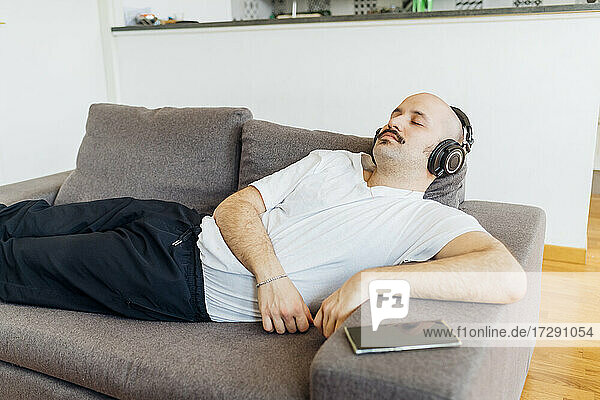 Glatzköpfiger Mann mit geschlossenen Augen hört Musik über Kopfhörer  während er sich zu Hause auf dem Sofa entspannt