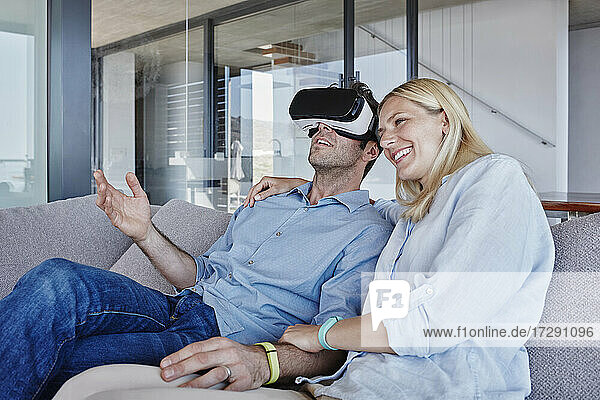 Lächelnde Frau sitzt neben einem Mann  der einen Virtual-Reality-Simulator im Wohnzimmer trägt