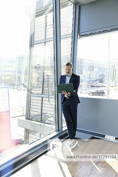 Geschäftsmann arbeitet am Laptop  während er sich im Büro ans Fenster lehnt