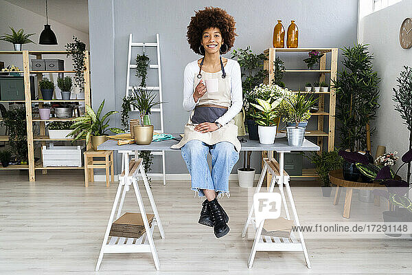 Weibliche Ladenbesitzerin mit Kaffee in der Hand  während sie auf einem Tisch in einem Pflanzengeschäft sitzt