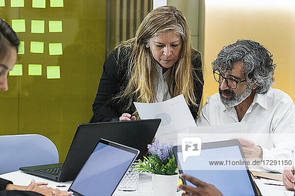 Weibliche Fachkraft  die mit einem älteren Geschäftsmann über ein Dokument diskutiert  während Kollegen im Büro am Laptop arbeiten