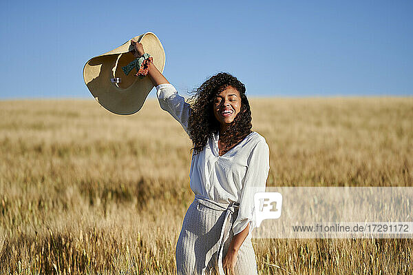 Fröhliche junge Frau mit lockigem Haar  die einen Hut hält  während sie auf einem Weizenfeld vor blauem Himmel steht