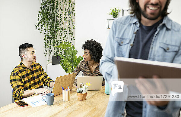 Männliche und weibliche Berufstätige mit Laptop diskutieren am Schreibtisch sitzend  während ein Kollege ein digitales Tablet im Büro benutzt