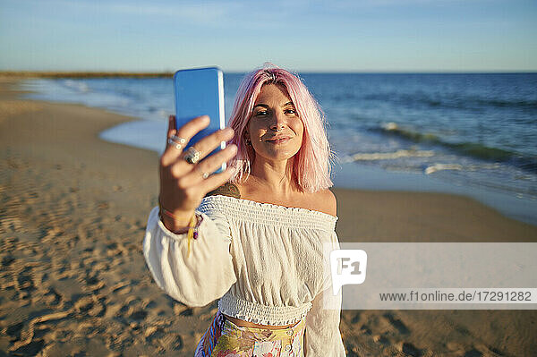 Junge Frau  die ein Selfie mit ihrem Smartphone macht  während sie am Strand bei Sonnenuntergang steht