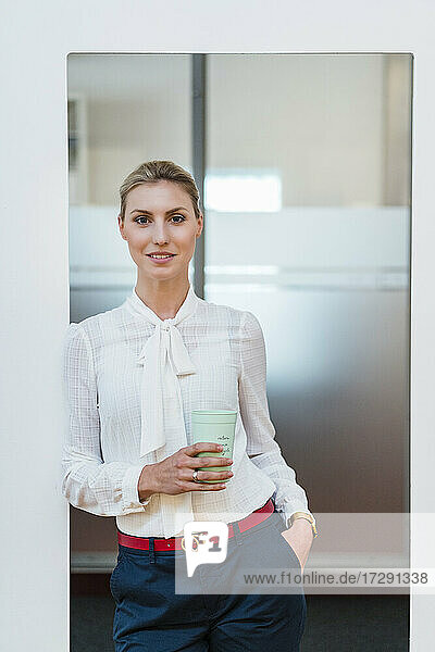 Schöne junge Geschäftsfrau steht mit Einweg-Kaffeebecher an der Tür im Büro