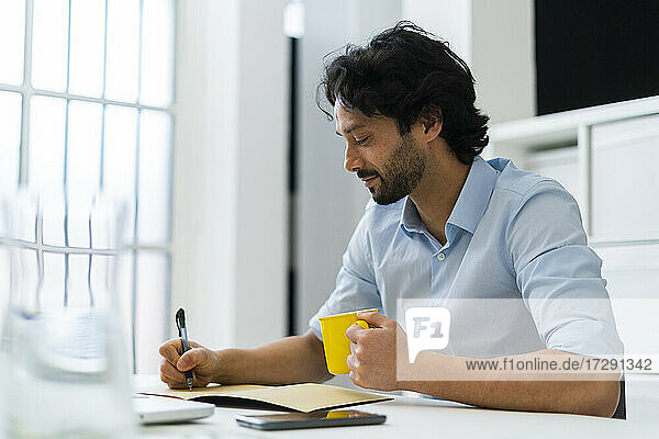 Male entrepreneur with coffee cup writing in book at office