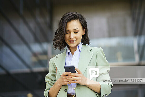 Lächelnde weibliche Fachkraft  die ein Smartphone in der Nähe eines Bürogebäudes benutzt