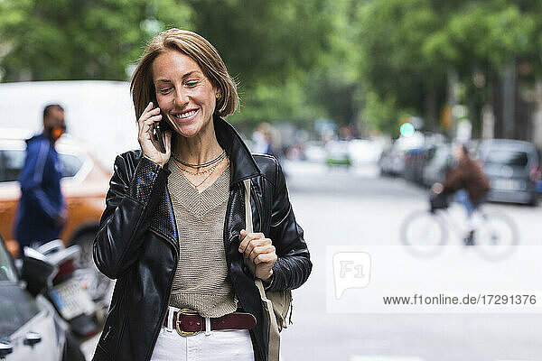 Lächelnde Frau mit Umhängetasche  die auf einer Straße in der Stadt mit einem Mobiltelefon spricht