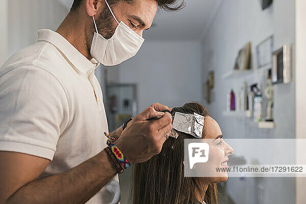 Männlicher Experte mit Gesichtsmaske beim Färben der Haare einer lächelnden Frau im Friseursalon