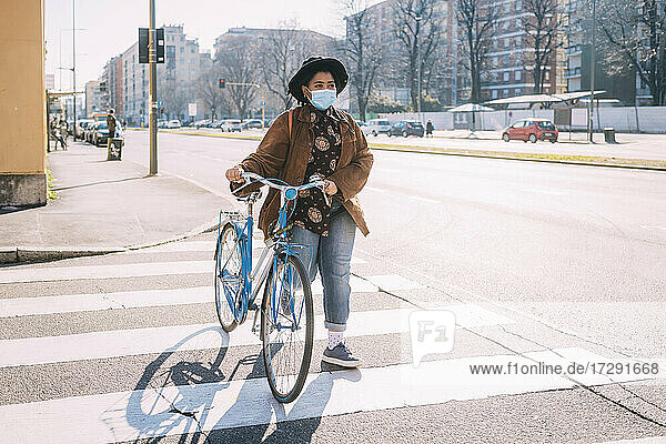 Junge Frau mit Gesichtsmaske und Hut geht mit dem Fahrrad auf einer Straße in der Stadt spazieren