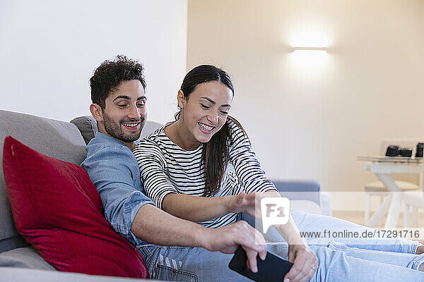 Glückliches Paar lachend und mit Handy spielend auf dem Sofa zu Hause
