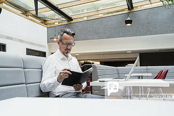 Businessman checking book while sitting by table in office lobby