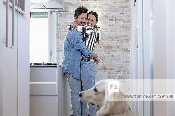 Lächelndes  sich umarmendes Paar mit Blick auf den Hund in der heimischen Küche zu Hause