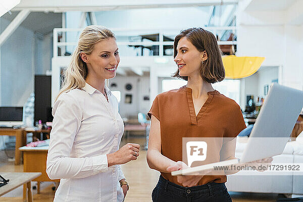 Lächelnde blonde Geschäftsfrau  die eine Kollegin ansieht  die einen Laptop hält  während sie in einem kreativen Büro diskutiert