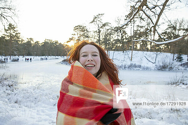 Fröhliche rothaarige Frau in Decke eingewickelt im Winter bei Sonnenuntergang