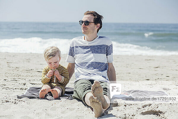Entspannter Mann  der wegschaut  während er neben seinem Sohn sitzt und einen Snack am Strand isst