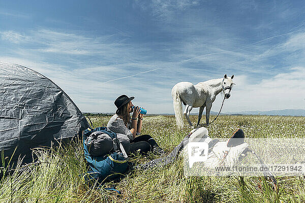 Junge Frau trinkt Wasser auf einer Wiese neben einem Pferd an einem sonnigen Tag
