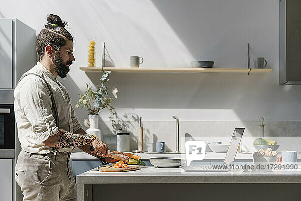 Bärtiger Mann schneidet Gemüse  während er in der Küche auf seinen Laptop schaut
