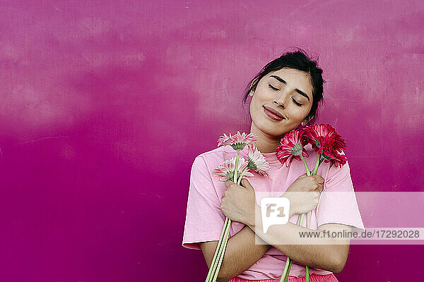 Junge Frau mit verschränkten Armen  die einen Strauß Gerbera-Gänseblümchen vor einer rosa Wand hält