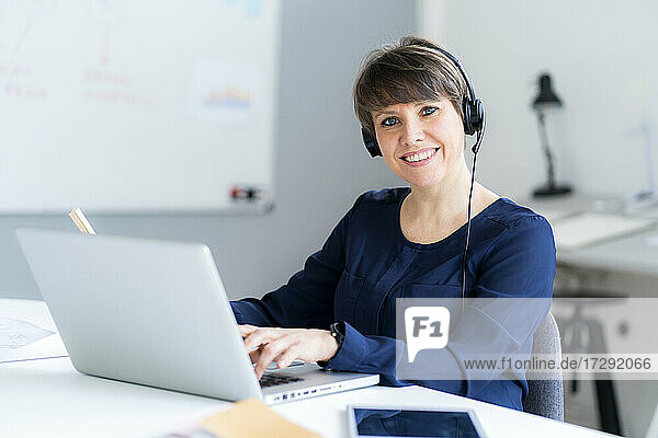 Lächelnde weibliche Fachkraft mit Headset sitzt mit Laptop am Schreibtisch im Büro