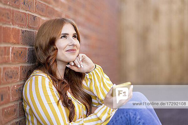 Lächelnde nachdenkliche Frau sitzt mit Smartphone an einer Backsteinmauer