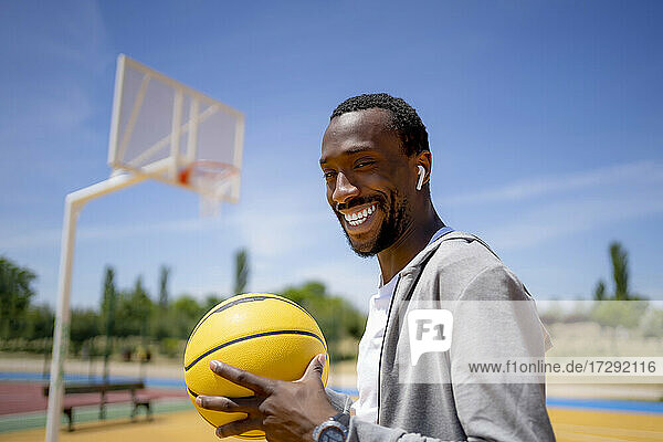 Lächelnder junger Mann mit drahtlosen In-Ear-Kopfhörern  der an einem sonnigen Tag einen Basketball hält